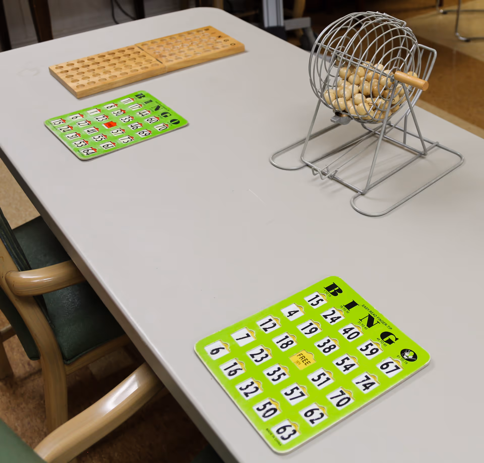 A table with two green bingo cards, a metal bingo cage with wooden balls, and a wooden bingo number holder. Two chairs with wooden arms and green cushions are partially visible around the table.