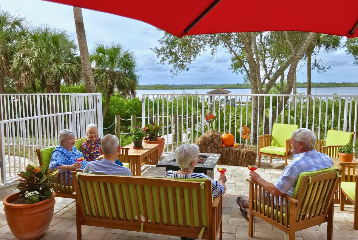 A group of five elderly people sitting on green cushioned wooden outdoor furniture under a red umbrella, enjoying drinks and conversation on a patio overlooking a body of water with palm trees and greenery in the background. There are potted plants and fall decorations including pumpkins and hay bales on the patio.