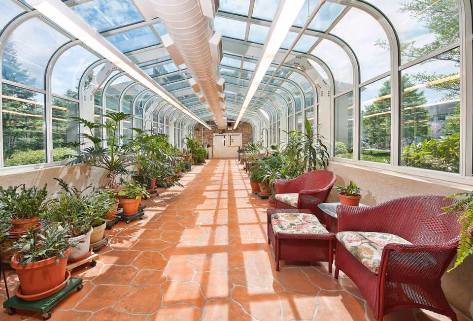 A bright sunroom with large curved glass windows and ceiling, terracotta tile floor, and numerous potted plants along the walls. On the right side, there are red wicker chairs and a small table with floral cushions. The sunroom leads to a double door at the far end.