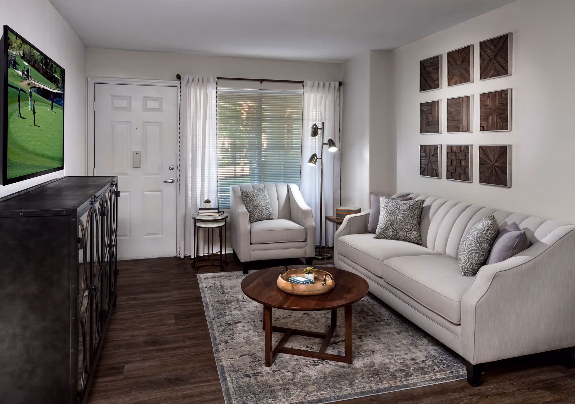 A modern living room with a light gray sofa and matching armchair, both adorned with patterned and solid pillows. A round wooden coffee table with a decorative tray sits on a patterned rug. Behind the armchair is a window with blinds and white curtains. On the wall above the sofa are nine square wooden art pieces arranged in a grid. A flat-screen TV is mounted on the wall opposite the sofa, and a dark cabinet is positioned below the TV. The room has wood flooring and a white door near the TV.