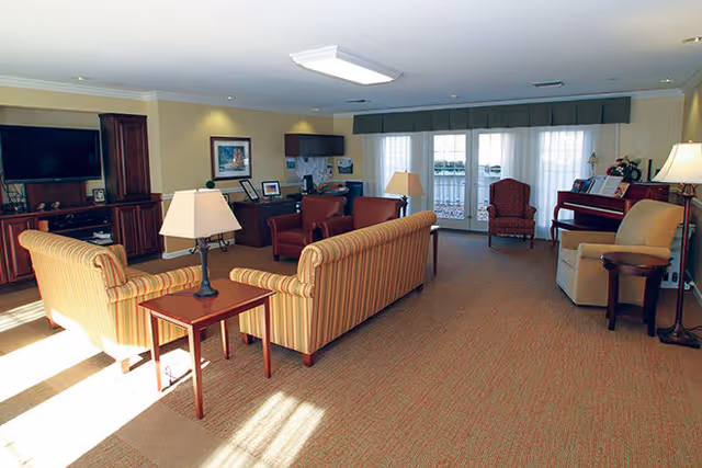 A spacious living room with beige carpet and cream-colored walls, featuring two striped sofas facing a wooden entertainment center with a flat-screen TV. There are two armchairs near a set of French doors with sheer curtains, a piano with a lamp on top, and several table lamps providing warm lighting. The room has a cozy and inviting atmosphere.