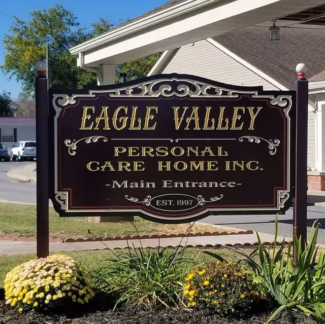 A decorative sign for Eagle Valley Personal Care Home Inc. at the main entrance, surrounded by flowers and greenery, with a building and parked cars visible in the background.