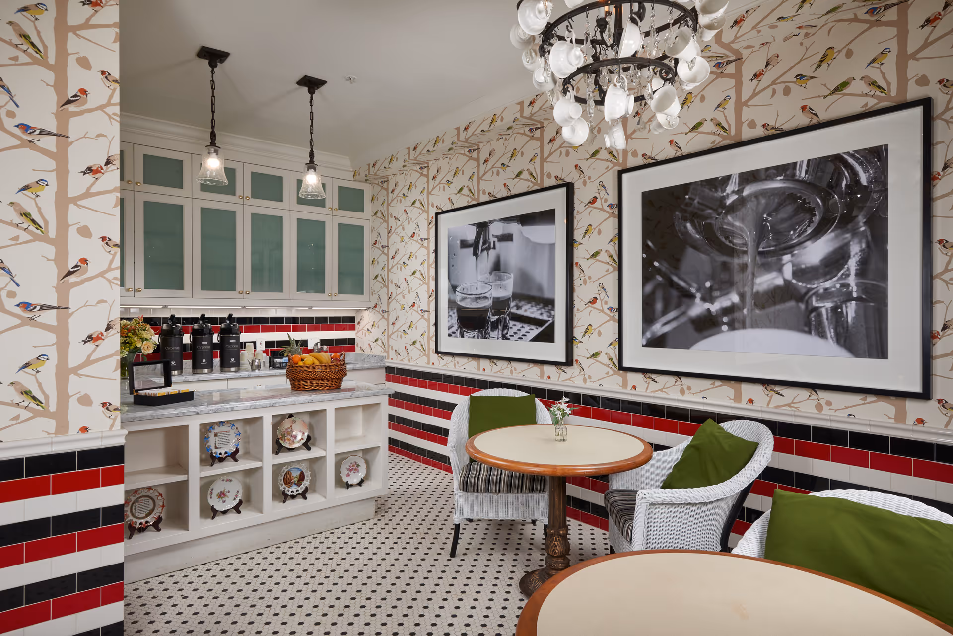 A cozy dining area with two round tables and white wicker chairs with green cushions. The walls are decorated with wallpaper featuring birds on branches and two large black and white framed photographs of coffee being poured. The lower half of the walls has a red, black, and white tiled pattern. There is a countertop with cabinets above and below, holding coffee dispensers and a basket of fruit. The floor has a black and white mosaic tile pattern, and two pendant lights hang from the ceiling.