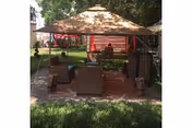 Outdoor seating area with a canopy tent providing shade over several chairs and a small table, set on a paved surface with grass surrounding it and trees in the background.