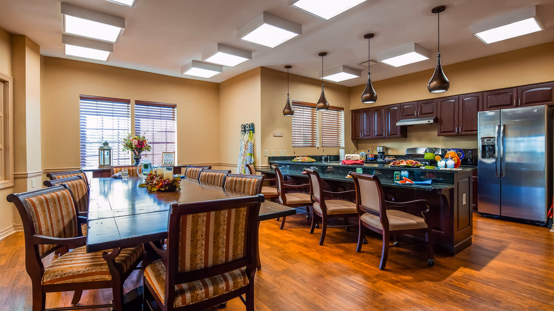 A spacious kitchen and dining area with wooden floors and beige walls. The dining area features a large wooden table surrounded by cushioned chairs with striped upholstery. The kitchen has dark wood cabinets, a granite countertop island with several chairs, stainless steel refrigerator, and modern pendant lights hanging from the ceiling. Two windows with blinds allow natural light into the room.