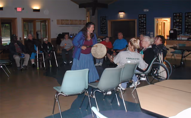 A group of elderly people seated in a semi-circle inside a common room, with a woman standing in the center playing a drum. The room has chairs, tables, and soft lighting, creating a communal and engaging atmosphere.