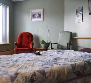 A cozy room with a bed covered in a patterned quilt in the foreground, a red cushioned armchair and a green chair near the wall, a small table with a potted plant between the chairs, a framed picture on the wall, and a calendar hanging on another wall.