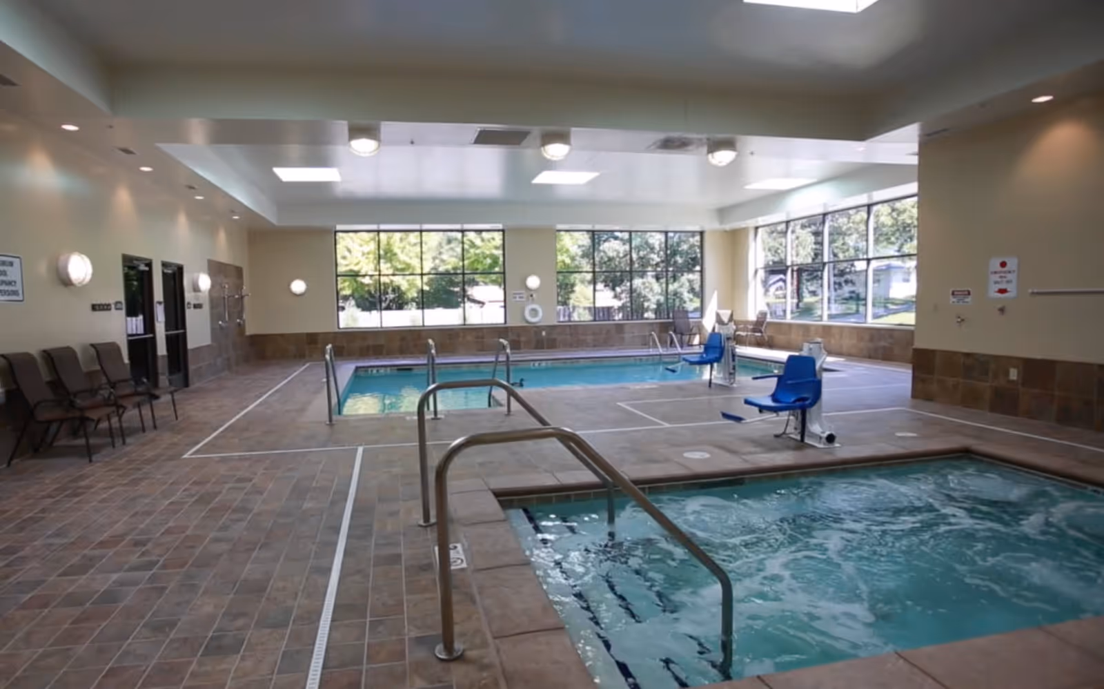 Indoor pool area with a hot tub in the foreground and a swimming pool in the background. The room has large windows letting in natural light, tiled floors, metal handrails for pool access, and blue pool lifts for accessibility. Several chairs are lined up along the left wall.