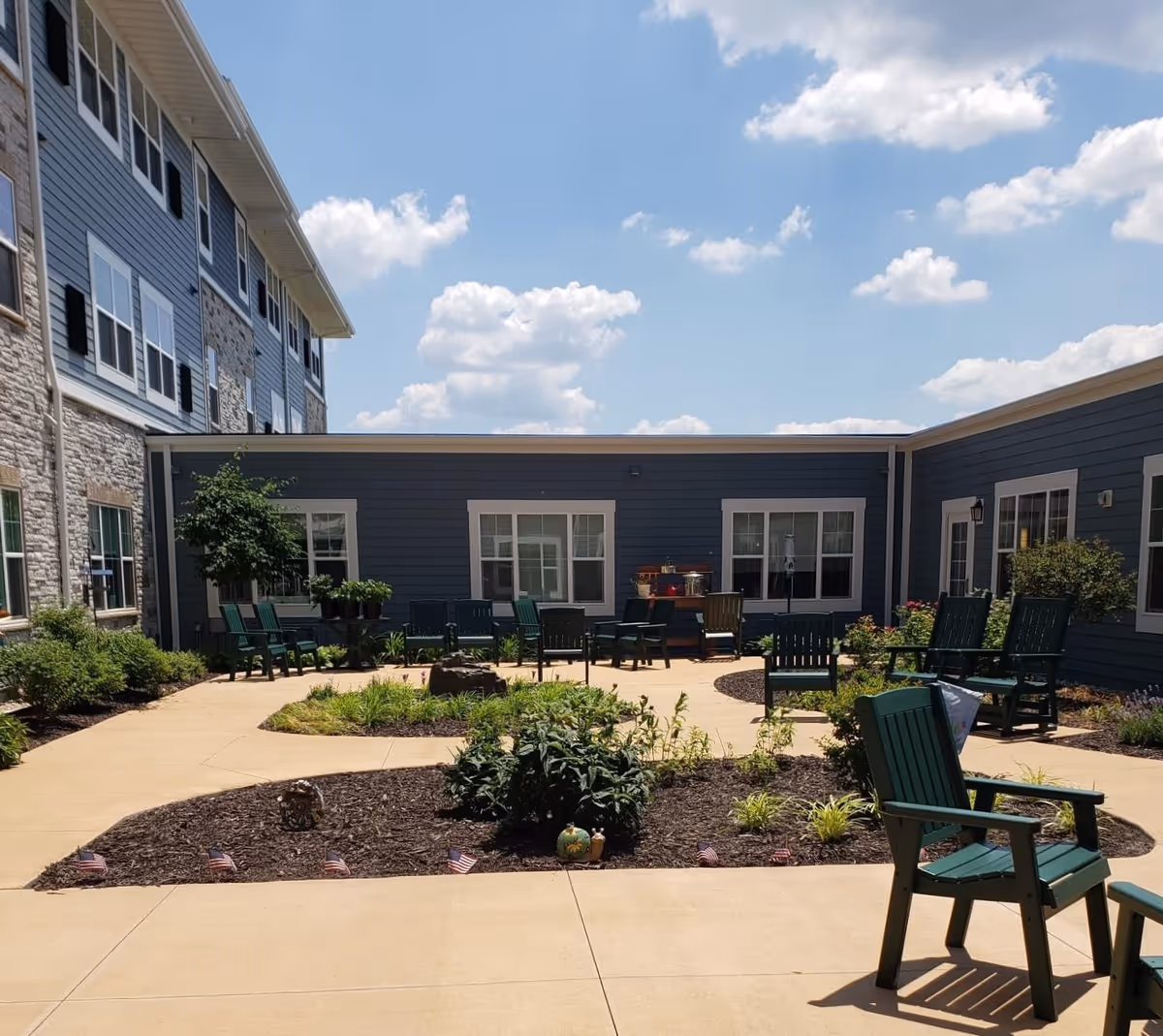 Outdoor courtyard area at Clarendale of St. Peters with green chairs arranged around a circular garden bed. The building exterior is blue with white trim and multiple windows. The sky is partly cloudy.
