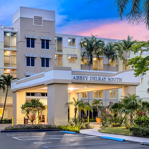 Exterior view of Abbey Delray South facility at dusk with a covered entrance, palm trees, and landscaped greenery in front of the building.