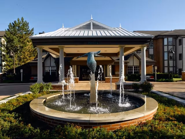 Front entrance of a senior living facility with a covered driveway supported by white columns. In front of the entrance is a circular fountain with water jets and a sculpture of a bird's tail. The building has a brick and beige exterior with multiple windows and a peaked roof.