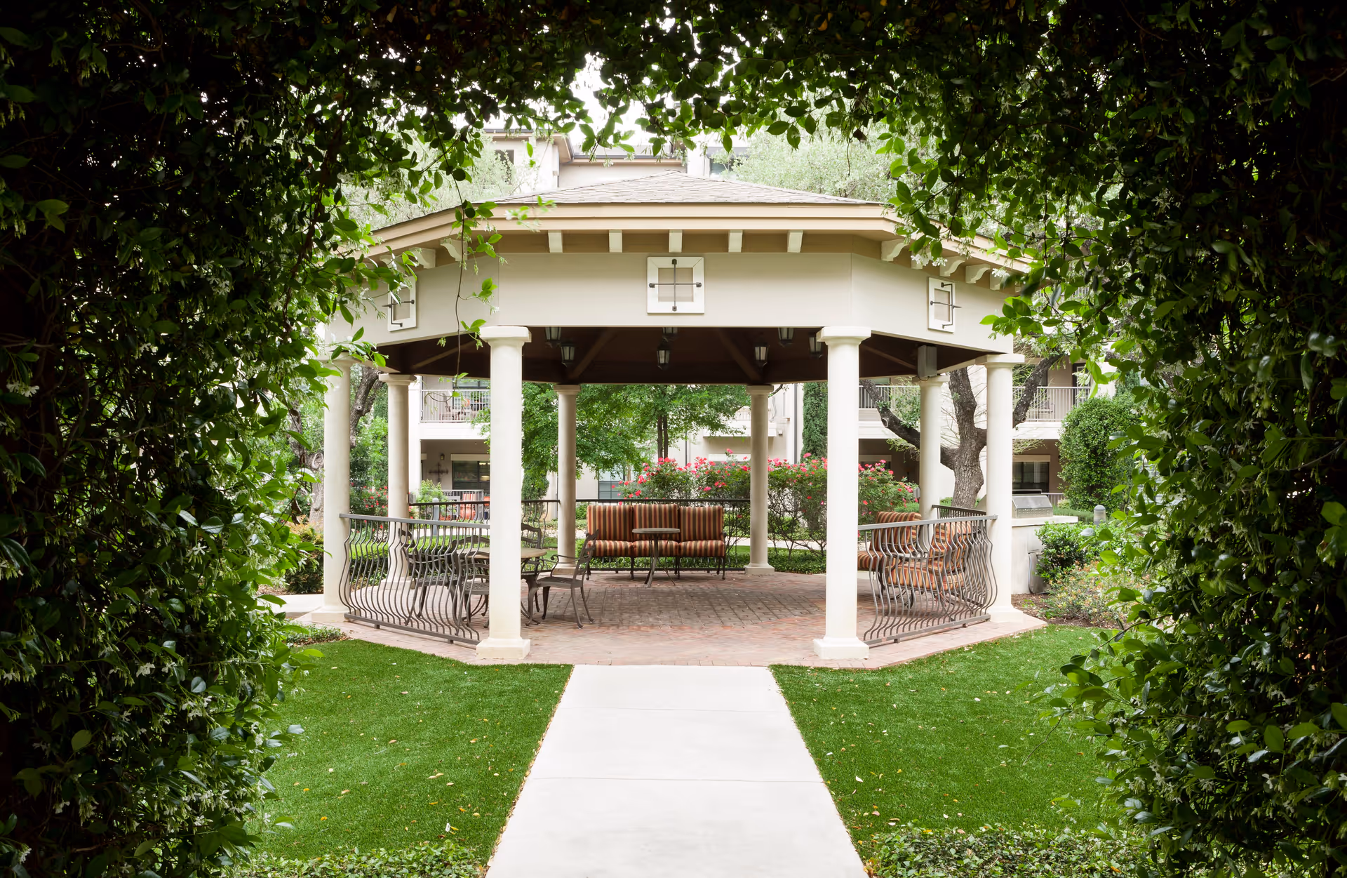 A covered outdoor gazebo with benches and tables in a landscaped courtyard framed by leafy greenery.