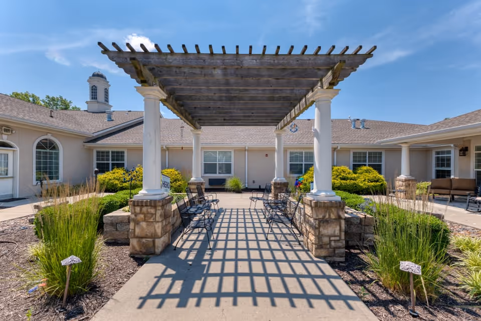 Outdoor courtyard area at Addington Place of Prairie Village featuring a wooden pergola with white columns and stone bases, surrounded by benches and landscaped greenery under a clear blue sky.