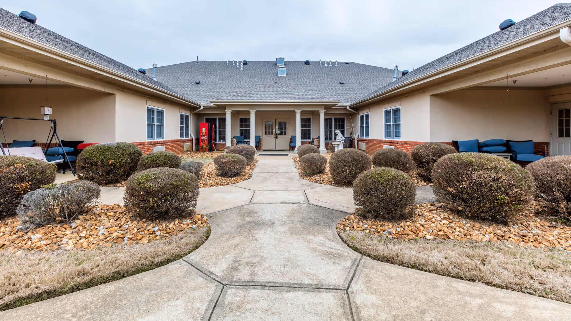 Outdoor courtyard area of a senior living facility with trimmed bushes, stone landscaping, and a concrete walkway leading to the entrance of a beige building with a gray roof. There are seating areas with blue cushions on both sides under covered walkways.