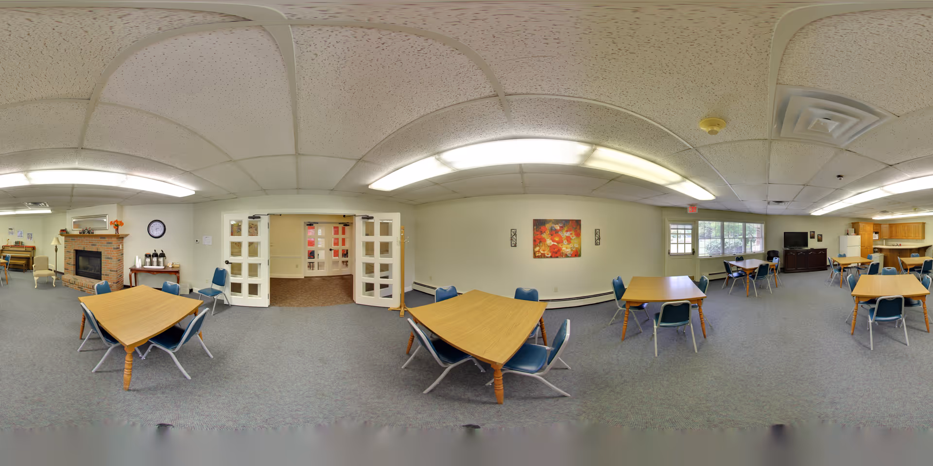 A spacious common area in Grace Village Retirement Community featuring multiple wooden tables with blue chairs arranged around them. The room has a carpeted floor, a brick fireplace with a mantle decorated with a clock and flower vase, and a wall with a colorful floral painting. There are double glass doors leading to another room, large windows letting in natural light, and a kitchenette area with cabinets and appliances in the background.