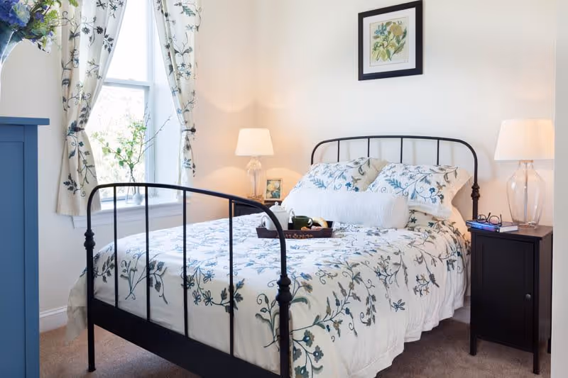 A cozy bedroom with a black metal bed frame, floral patterned bedding, and matching curtains. There are two bedside tables with lamps, a framed botanical print on the wall, and a tray with a cup and teapot on the bed.