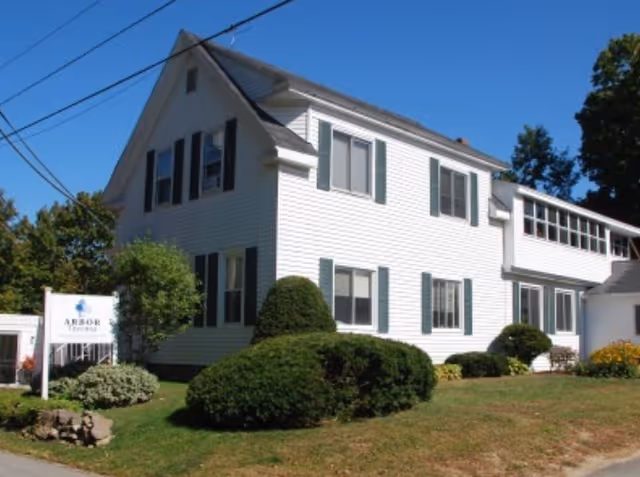 White two-story building with green shutters and a well-maintained lawn with bushes in front under a clear blue sky. A sign near the road reads 'Arbor Terrace'.
