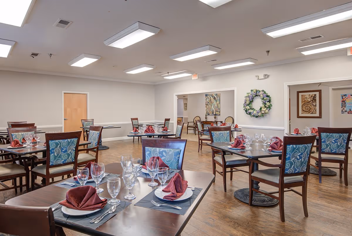 A senior living facility dining room with multiple tables set with plates, glasses, silverware, and folded burgundy napkins. The chairs have blue patterned upholstery, and the room features wood flooring, white walls, and ceiling lights. There is a floral wreath on one wall and framed artwork in the background.