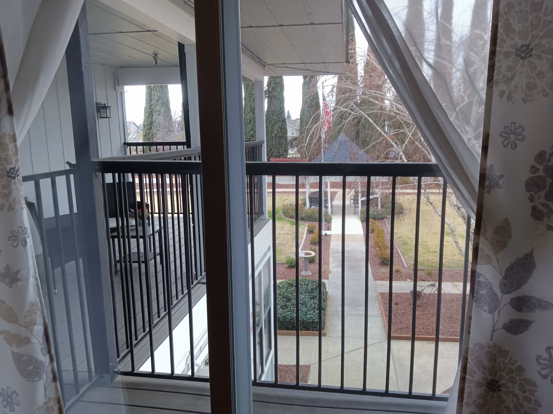 View through a sliding glass door with floral curtains, looking out onto a balcony with black railings and chairs. Beyond the balcony is a landscaped courtyard with a paved walkway, bushes, trees, and a gazebo.