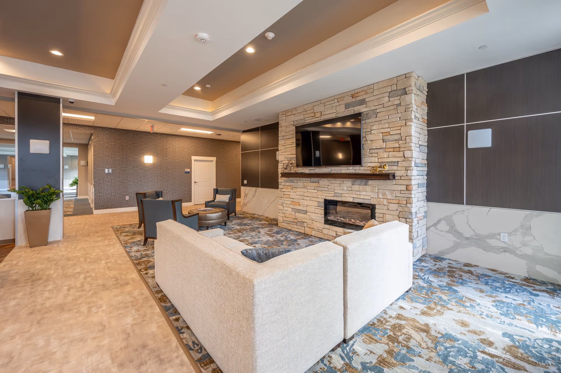 A modern and cozy living room area in a senior living facility featuring a beige sectional sofa, two dark armchairs, a round ottoman, and a stone fireplace with a mounted flat-screen TV above it. The room has a patterned carpet with blue and brown tones, recessed lighting in the ceiling, and decorative wall panels.