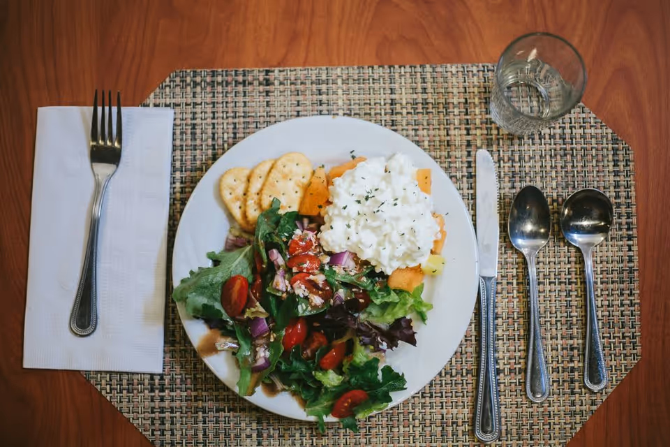 A plate with a fresh salad including leafy greens, cherry tomatoes, diced red onions, and a serving of cottage cheese with crackers on the side. The plate is placed on a woven placemat on a wooden table. To the left of the plate is a fork on a folded white napkin, and to the right are a knife and two spoons. A glass of water is also visible at the top right corner of the placemat.