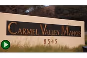 A large stone sign with the text 'Carmel Valley Manor' and the number '8545' displayed below it, set outdoors with some grass and trees in the background.