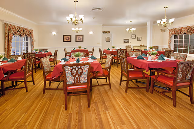 A dining room with multiple tables covered in red tablecloths, each set with green folded napkins, white plates, and silverware. The chairs have wooden frames with floral upholstery and red cushions. The room has wooden flooring, chandeliers hanging from the ceiling, and windows with floral curtains.