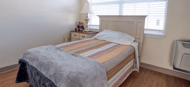 Single-occupancy bedroom with a made twin bed, wooden headboard, nightstand with a lamp and teddy bear, window blinds, and a wall air-conditioning unit.