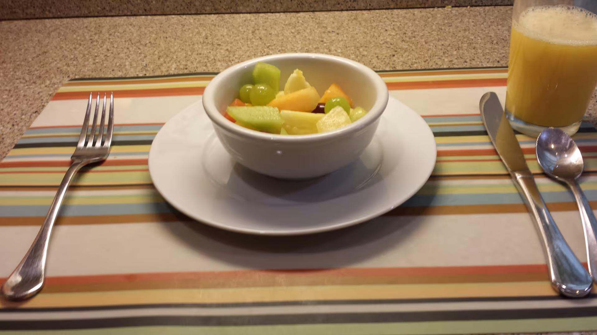 A bowl of mixed fruit including grapes, melon, and pineapple on a white plate, placed on a striped placemat. To the left of the plate is a fork, and to the right are a knife, spoon, and a glass of orange juice.