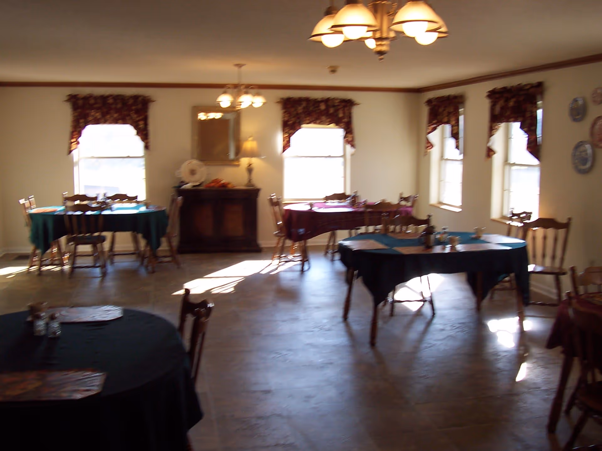 A dining room with several round and rectangular tables covered with dark tablecloths. Each table is surrounded by wooden chairs. The room has multiple windows with floral valances, allowing natural light to enter. There are chandeliers hanging from the ceiling and a sideboard with a lamp and decorative items against one wall.