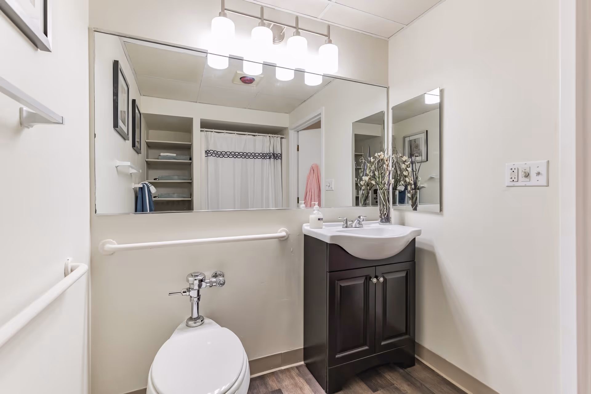 A clean and modern bathroom featuring a white toilet with a flush valve, a dark wood vanity with a white sink, a large mirror above the sink, and a smaller mirror on the adjacent wall. There is a light fixture with four lights above the large mirror, a white shower curtain with a decorative border, and grab bars on the walls for safety. The floor has a wood-like finish, and there are shelves with towels visible in the reflection.