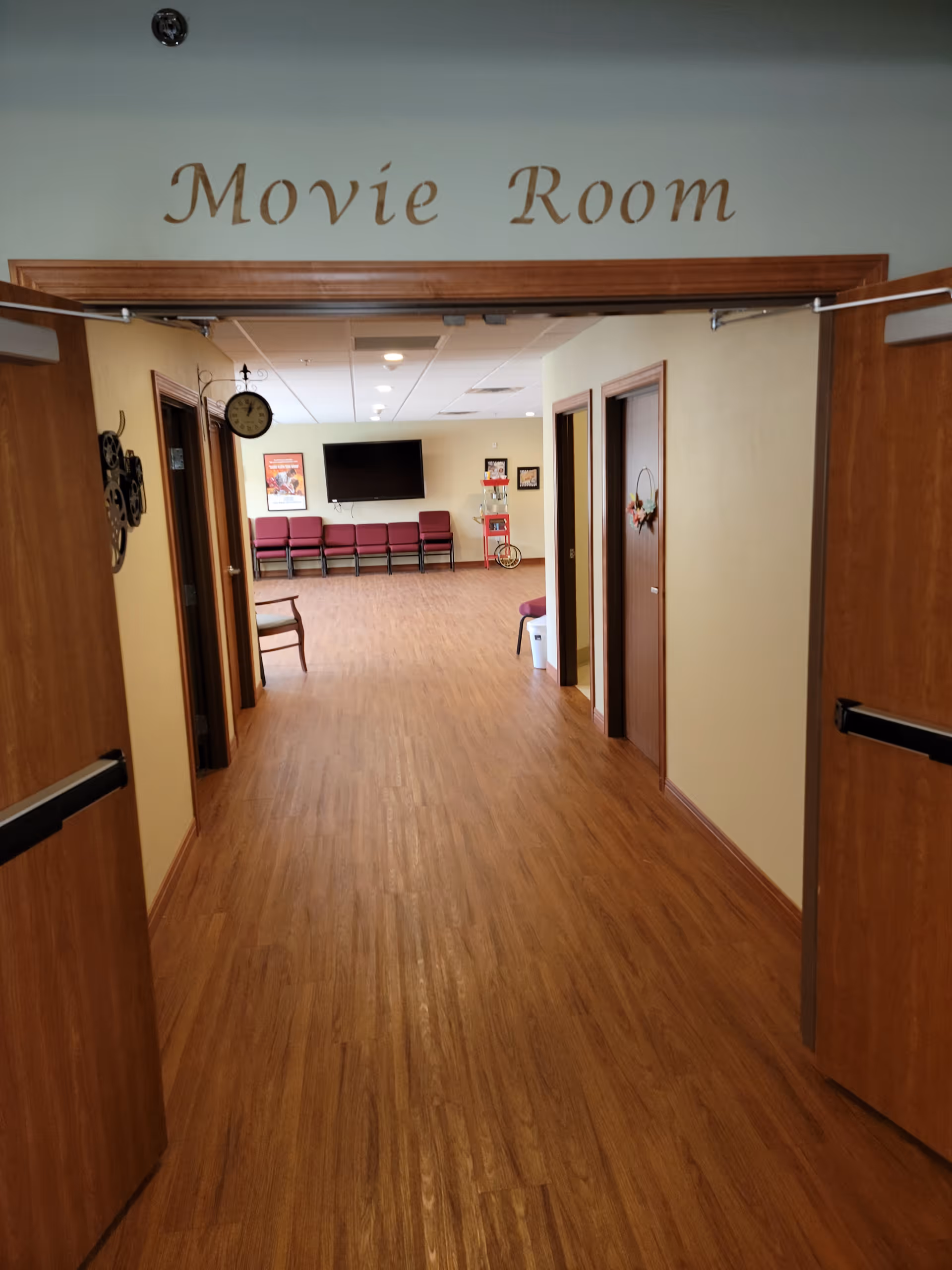View through open double doors into a movie room with wooden floors, beige walls, and a row of maroon chairs along the far wall beneath a mounted flat screen TV. There is a popcorn machine and framed pictures on the walls. The words 'Movie Room' are written above the doorway.