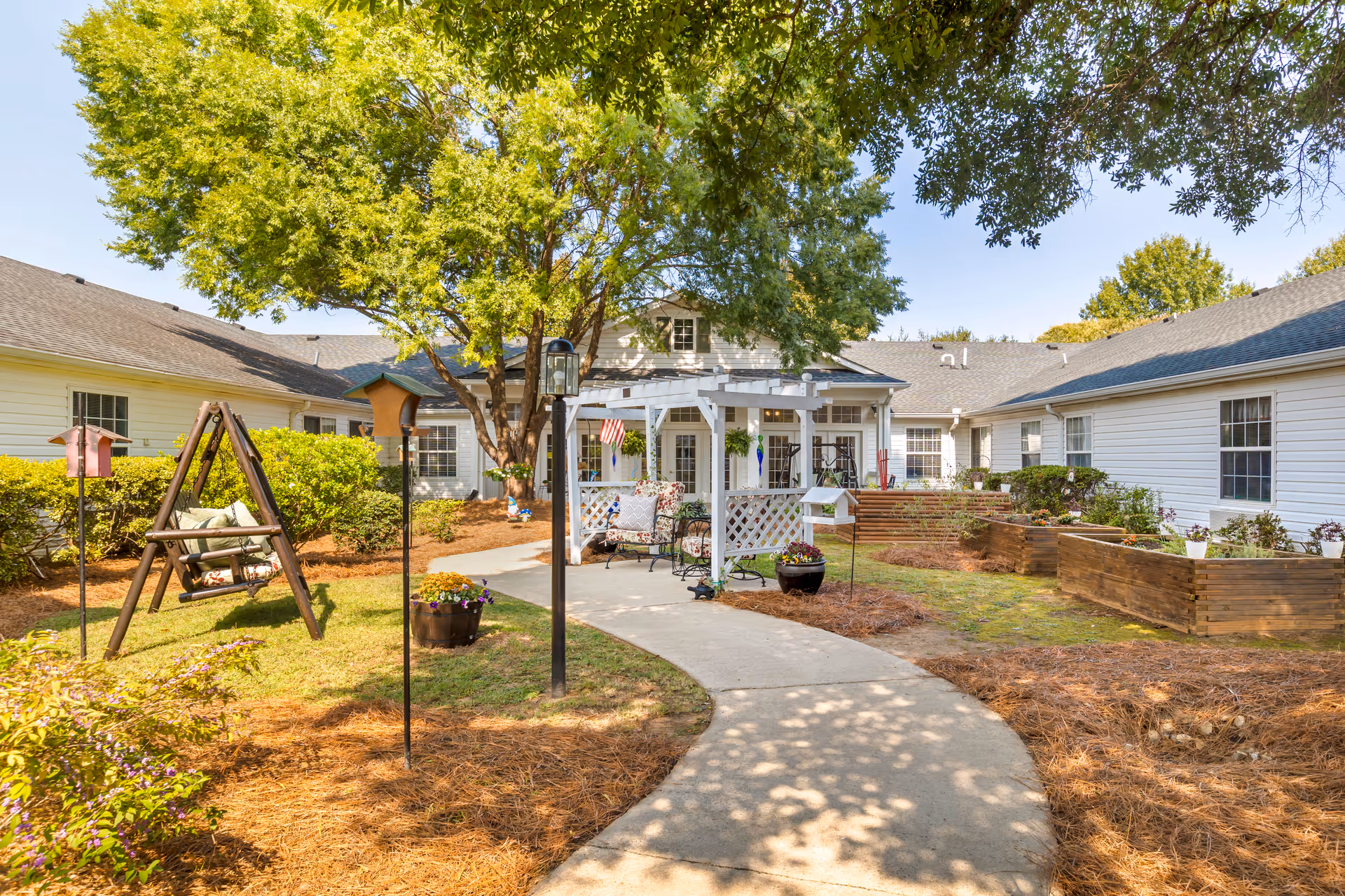 Outdoor courtyard area at Brookdale Brushy Creek featuring a curved concrete pathway, a wooden swing with cushions, a white pergola with seating underneath, raised garden beds, and a large tree providing shade. The courtyard is surrounded by a single-story building with white siding and multiple windows.