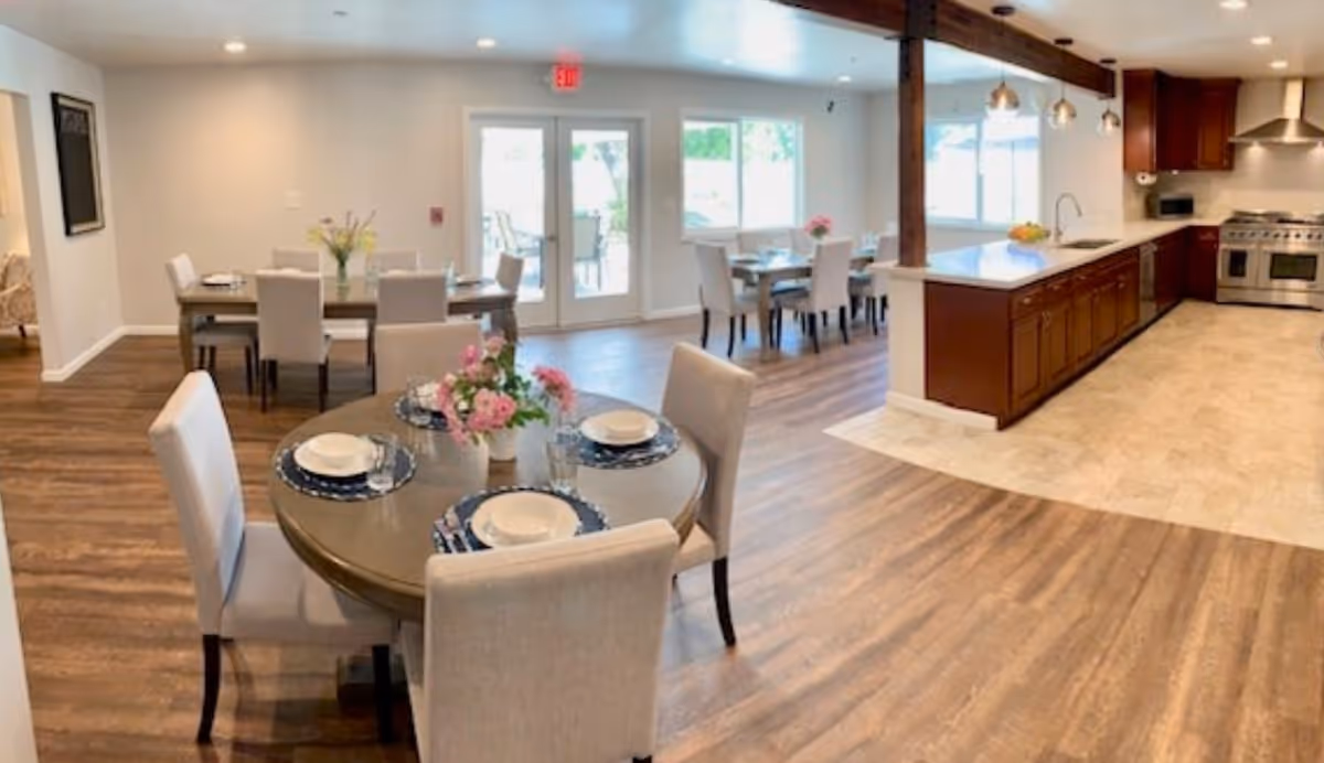A bright and spacious dining area with multiple tables set with plates, bowls, and glasses. The room features wood flooring and large windows letting in natural light. To the right is a modern kitchen with wooden cabinets, a large countertop, and stainless steel appliances including a stove and range hood. There are pendant lights hanging above the kitchen counter.