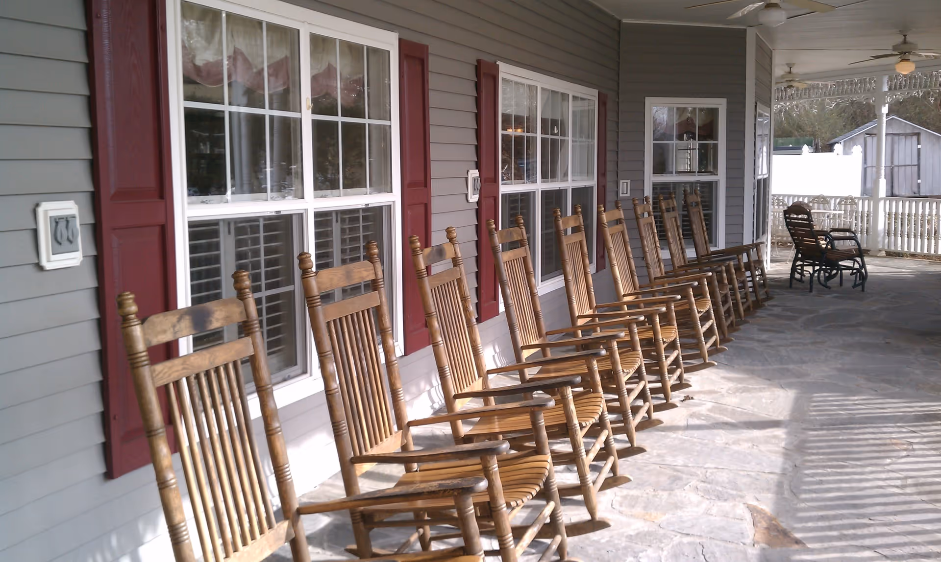 A long covered porch with a row of wooden rocking chairs lined up against the wall of a building with gray siding and red window shutters. The porch floor is made of stone, and there are two ceiling fans overhead. In the background, there are additional chairs and a white railing enclosing the porch.