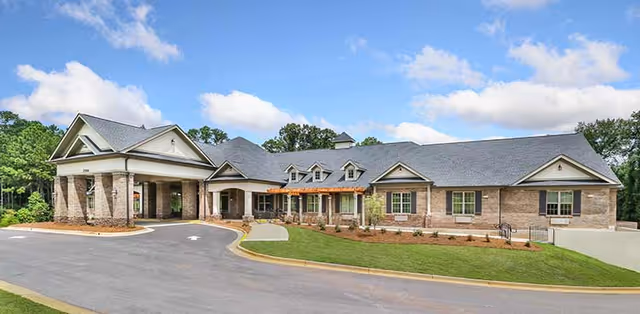Front exterior of a single-story brick senior living facility with a porte-cochere, landscaped lawn, and curved driveway under a blue sky.