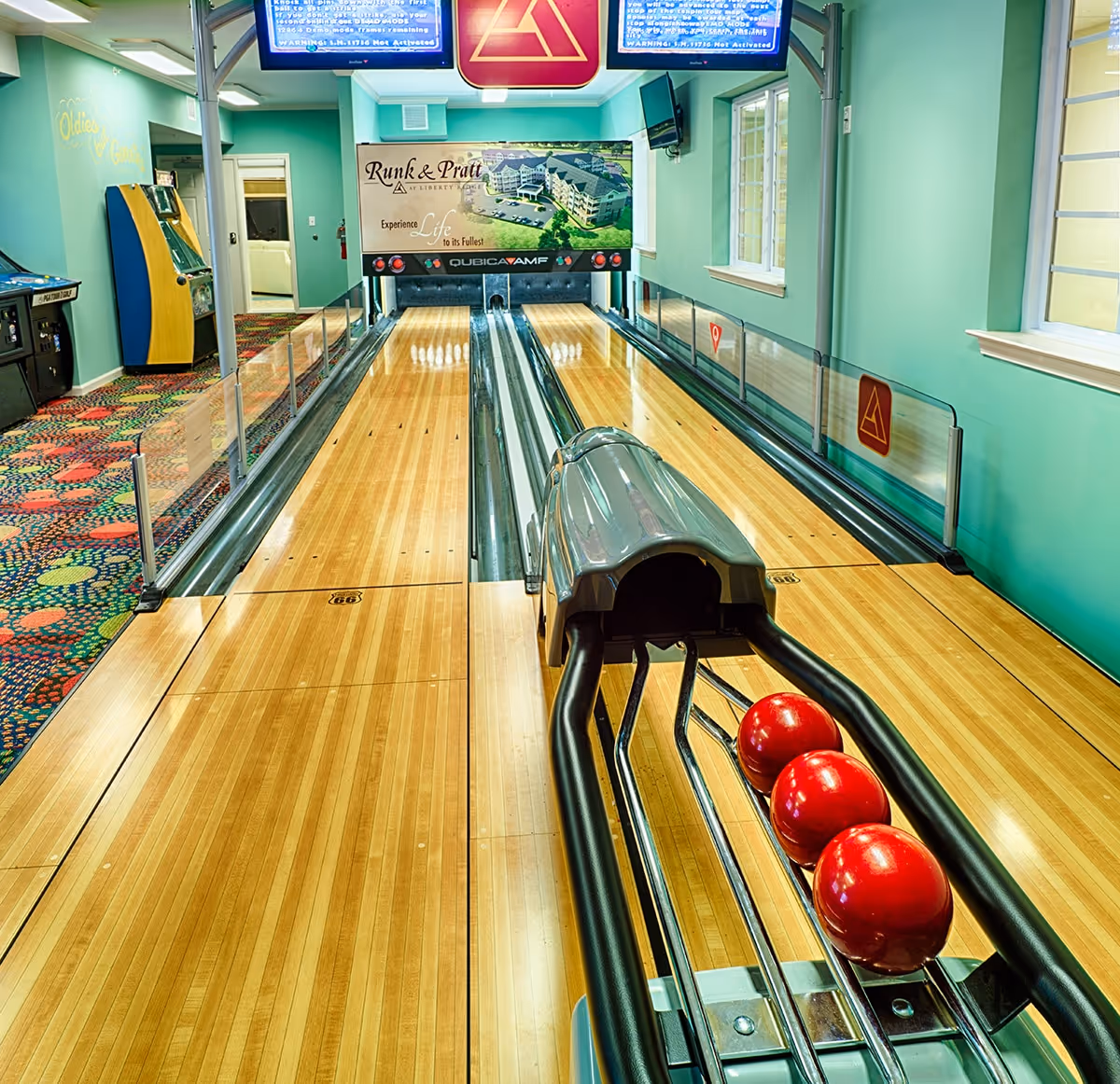 Indoor bowling alley with two lanes, three red bowling balls on the ball return rack, colorful patterned carpet on the left side, teal walls, windows on the right, and a sign at the end of the lanes that reads 'Runk & Pratt at Liberty Ridge Experience Life to its Fullest'.