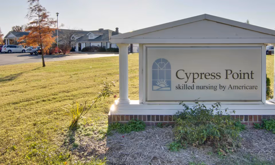 Outdoor view of the Cypress Point skilled nursing facility sign with a building and parking lot in the background, surrounded by grass and trees with autumn foliage.