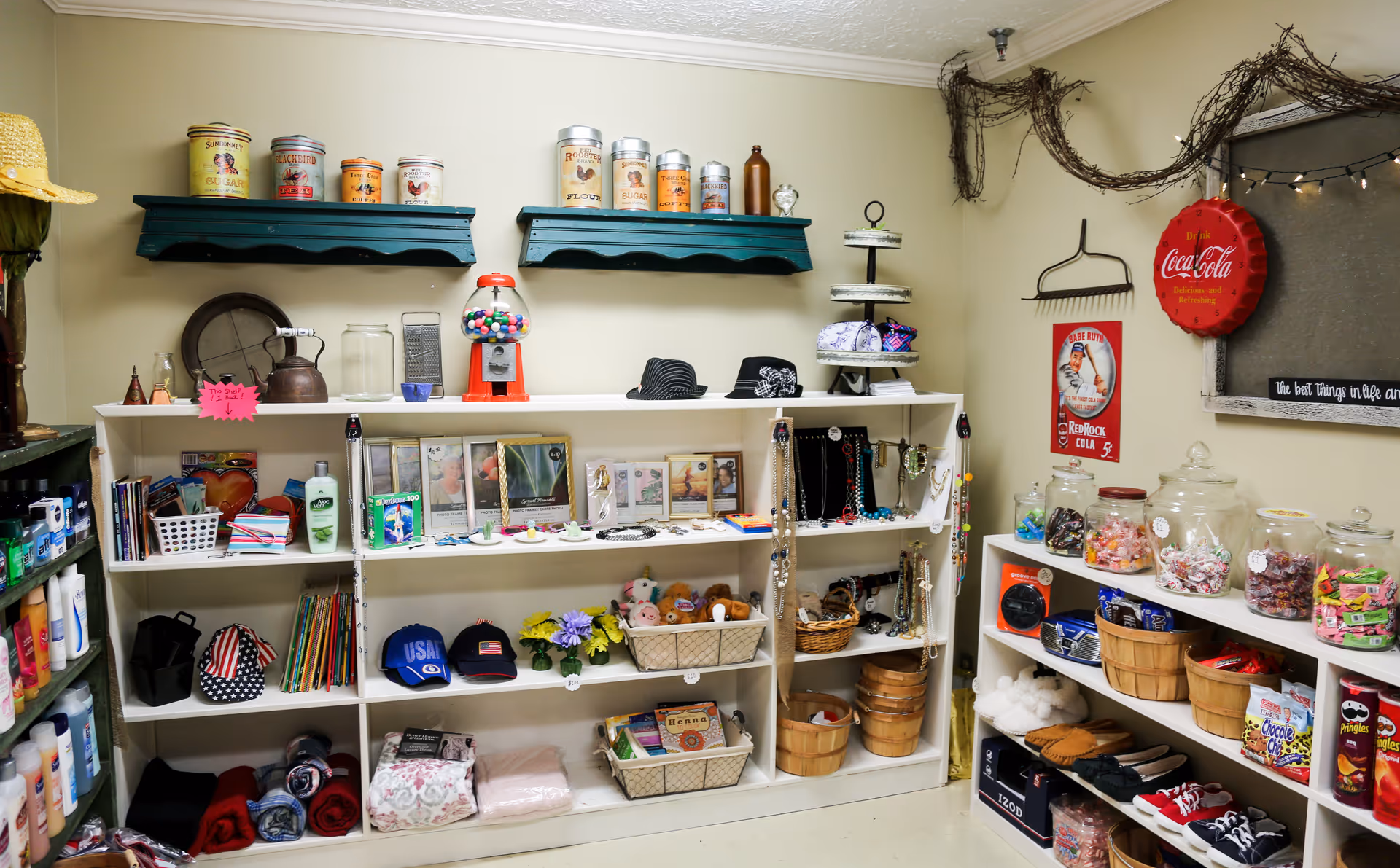 Small shop-like interior room with white shelving displaying hats, framed photos, jars of candy, toiletries, baskets and assorted gift items.