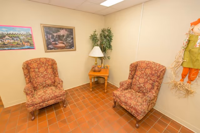 Two patterned armchairs flank a small wooden side table with a lamp in a tiled sitting area decorated with framed art and a potted plant.