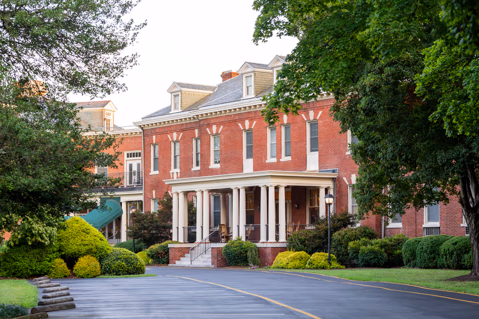 Exterior view of a large red brick building with white columns at the entrance, surrounded by green trees and bushes, with a paved driveway leading up to the entrance.