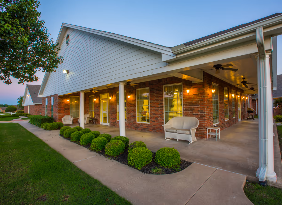 Exterior view of a brick building with a covered porch featuring white columns, outdoor seating, and ceiling fans. The porch is lit with warm wall-mounted lights, and there are neatly trimmed bushes along the sidewalk leading to the entrance. The sky is clear with a hint of dusk.