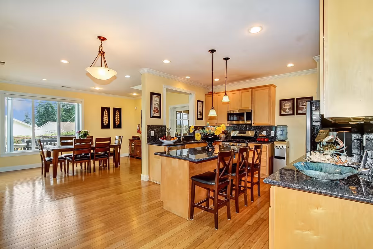 Bright and spacious kitchen and dining area with wooden floors and light yellow walls. The kitchen features light wood cabinets, black granite countertops, a kitchen island with three wooden bar stools, and stainless steel appliances. The dining area has a wooden table with six chairs and large windows letting in natural light. Decorative wall art and pendant lights add to the warm ambiance.