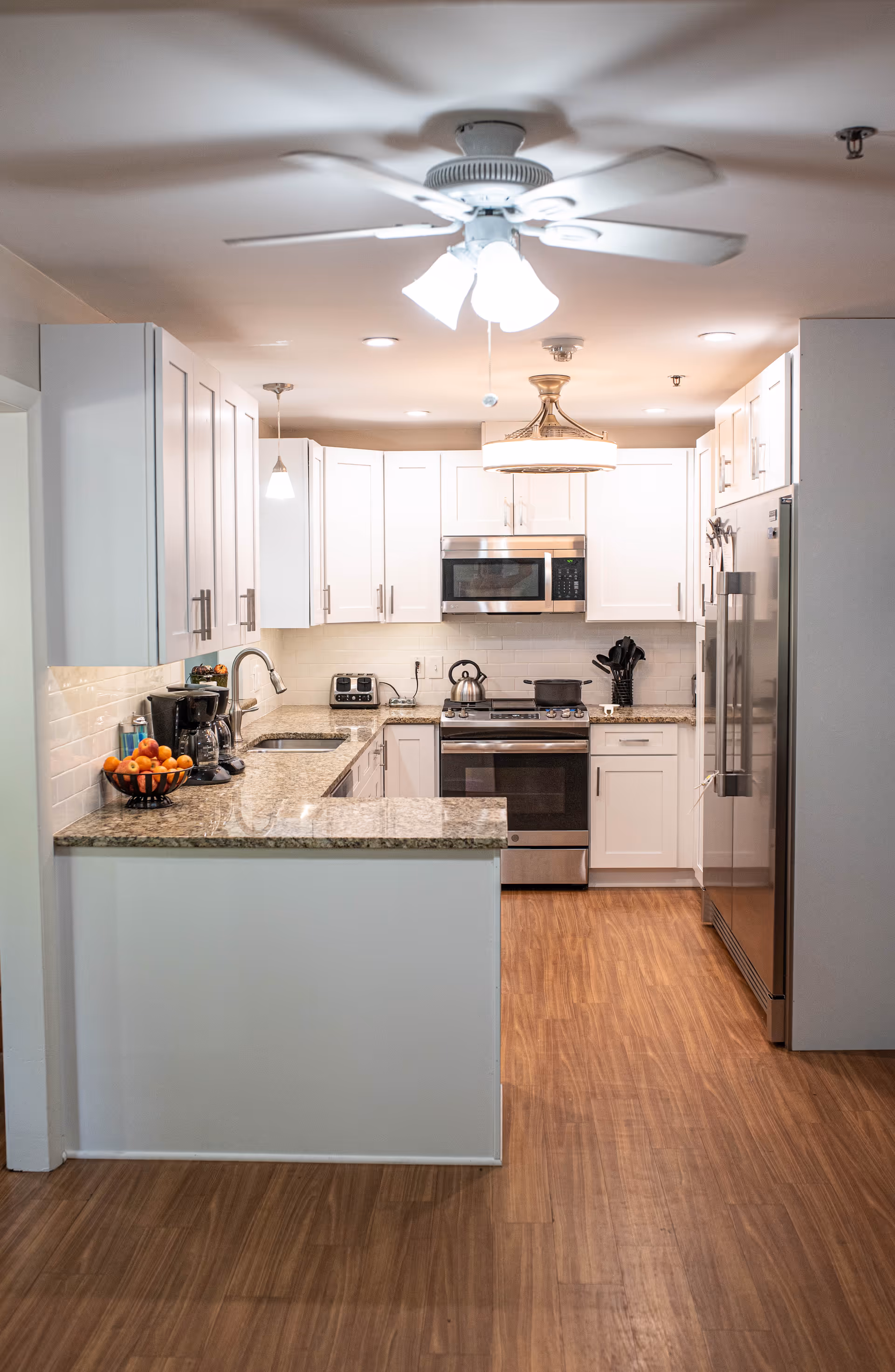 A modern kitchen with white cabinets, granite countertops, and stainless steel appliances including a refrigerator, oven, and microwave. The kitchen has a ceiling fan with lights, wooden flooring, and various kitchen items such as a coffee maker, toaster, and a bowl of oranges on the counter.
