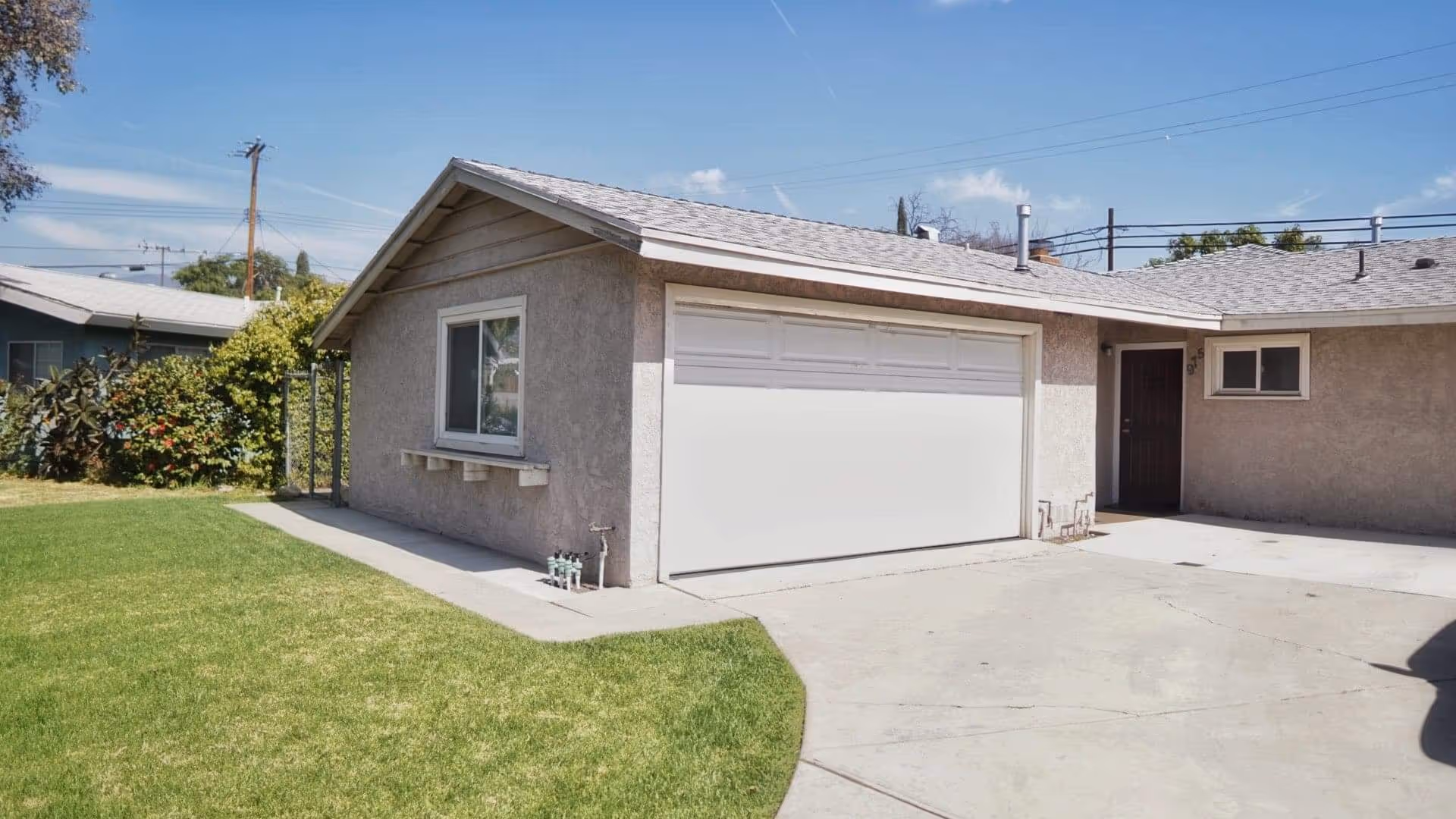 Exterior view of a single-story building with a gray stucco finish, a white garage door, a small window, and a dark entrance door. There is a concrete driveway and a green lawn adjacent to the building under a clear blue sky.