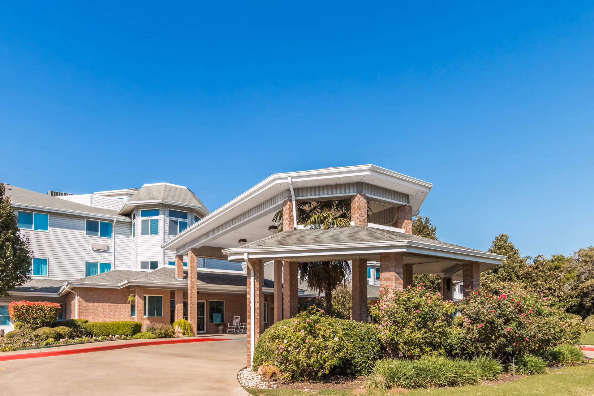 Exterior view of Asher Point Independent Living of Waco showing the entrance with a covered driveway supported by brick pillars, surrounded by well-maintained bushes and greenery under a clear blue sky.