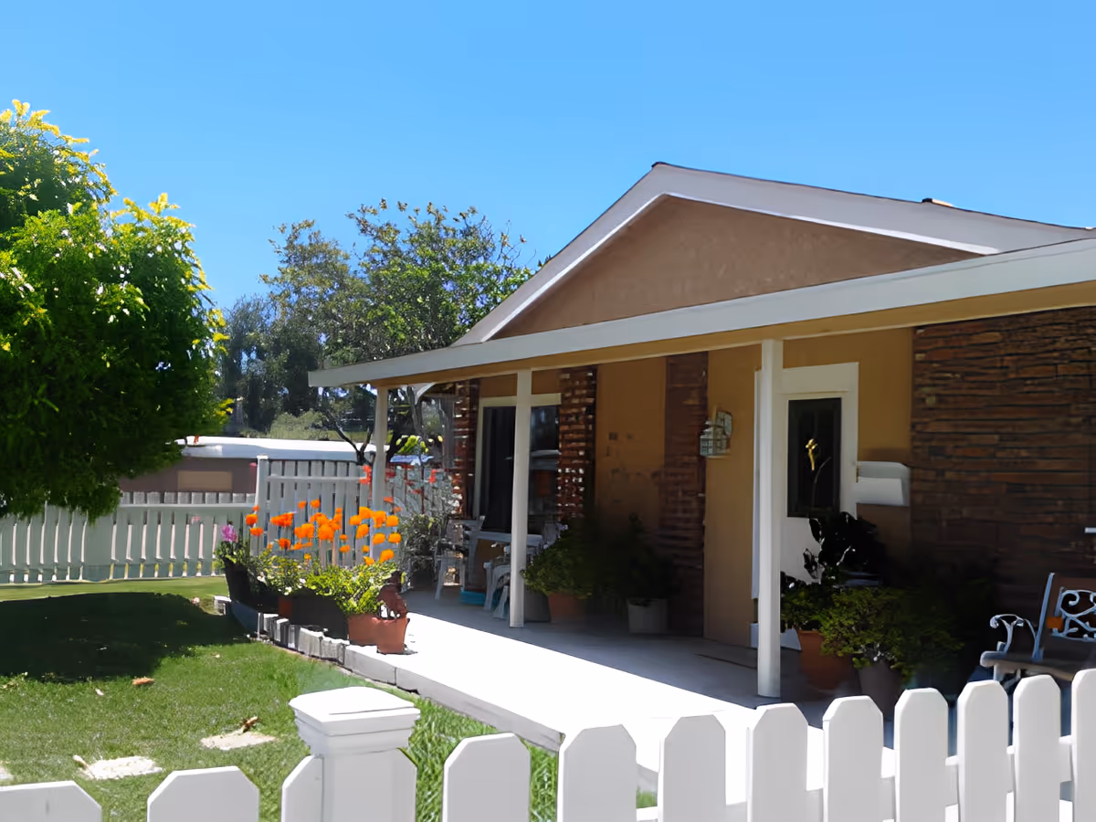 Front porch of a single-story building with a white picket fence, green lawn, potted plants, and orange flowers under a clear blue sky.