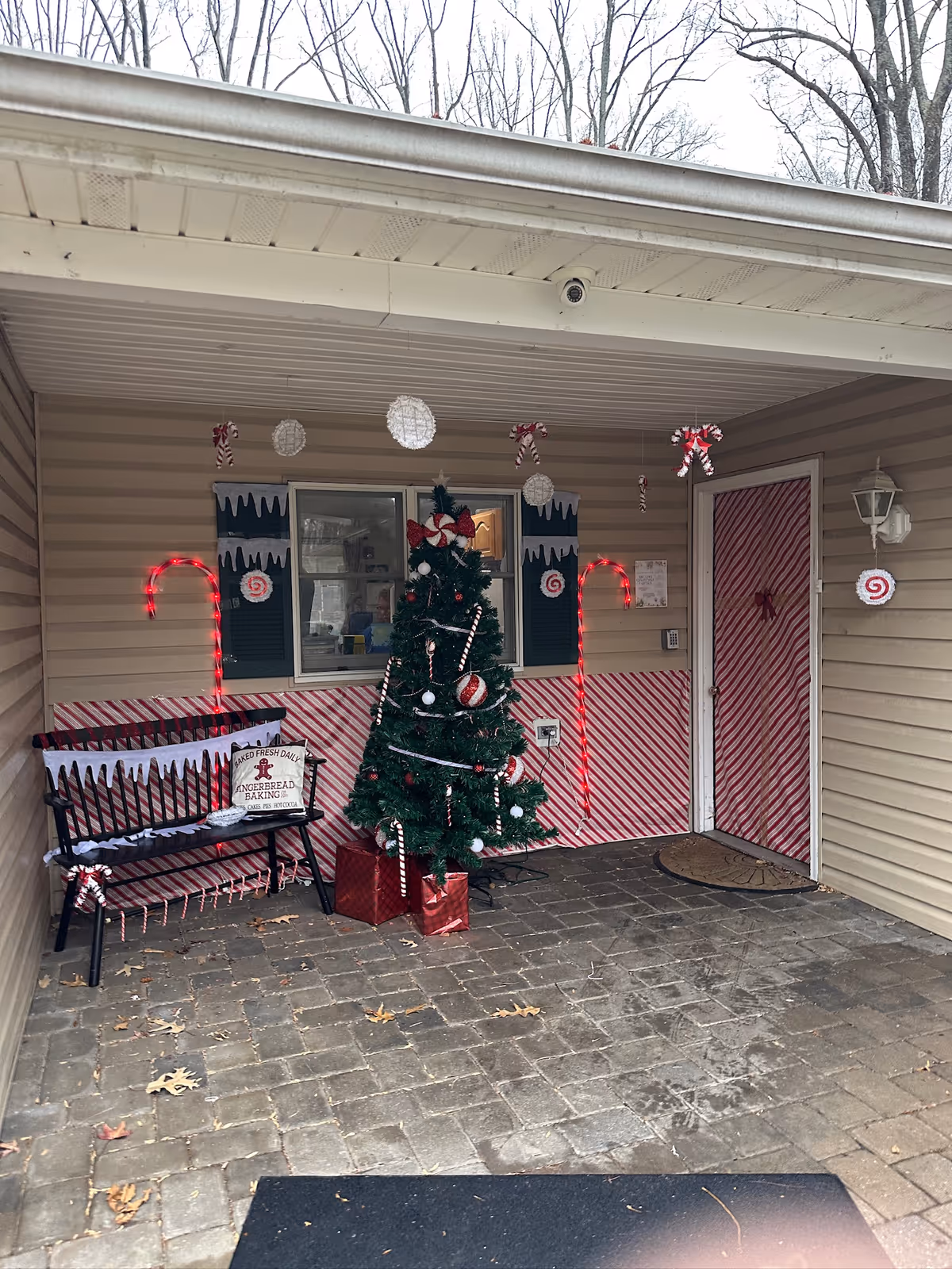 Covered porch area decorated for Christmas with a Christmas tree adorned with candy cane ornaments and red bows, two large red candy cane lights on either side of a bench, and red and white striped wrapping paper covering the wall and door. White hanging decorations and candy cane ornaments hang from the ceiling. A sign on the bench reads 'Fresh Baked Gingerbread Baking'.