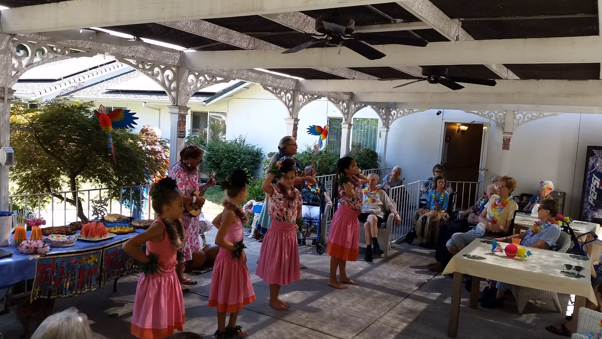 A group of young girls dressed in pink skirts and floral tops performing a hula dance under a covered patio. Two women are playing ukuleles nearby. Several elderly people are seated around tables and in chairs, watching the performance. The area is decorated with colorful tropical-themed decorations, including leis and paper parrots.