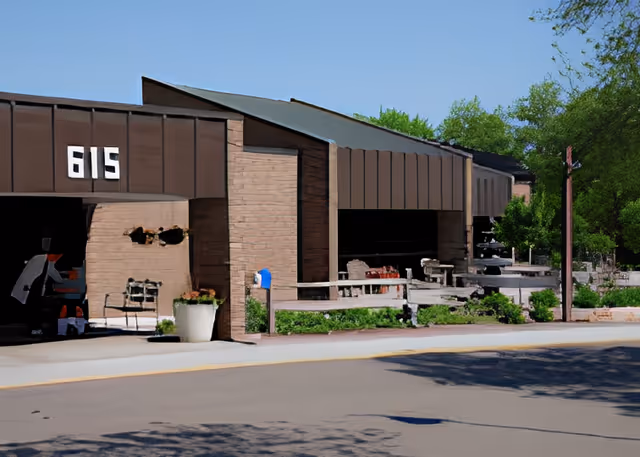 Exterior view of a building with the number 615 displayed prominently. The building has a brown facade with a covered entrance and some outdoor seating areas. There are trees and greenery surrounding the building under a clear blue sky.